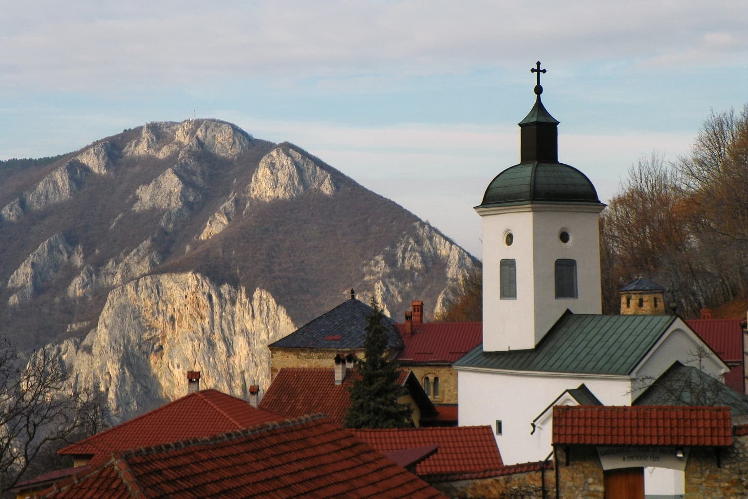 A monastery with a green-roofed tower and cross, red-tiled roofs, and a mountainous backdrop under a partly cloudy sky.