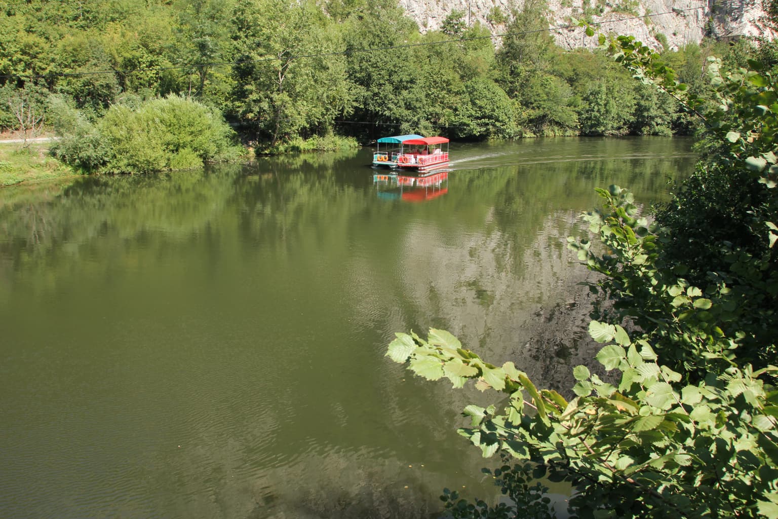A calm lake reflecting green trees and a colorful boat with a red and blue roof, surrounded by dense foliage