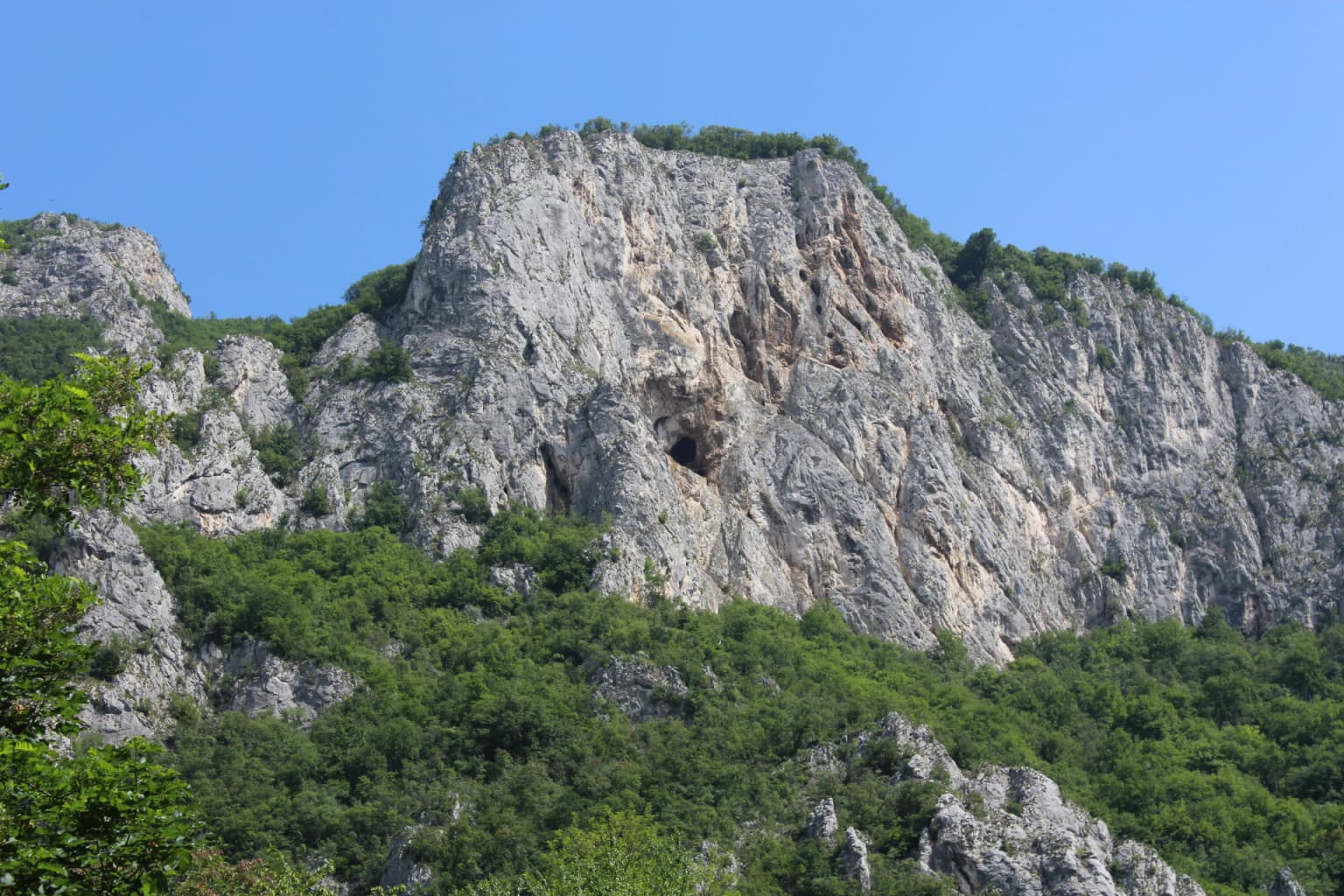 Rocky mountain with a cave opening, surrounded by dense green forest under clear blue sky