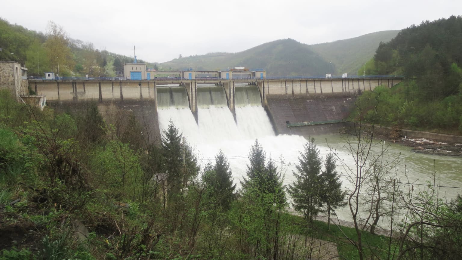 Large concrete dam with water flowing over spillway, surrounded by green vegetation and hills, with river downstream