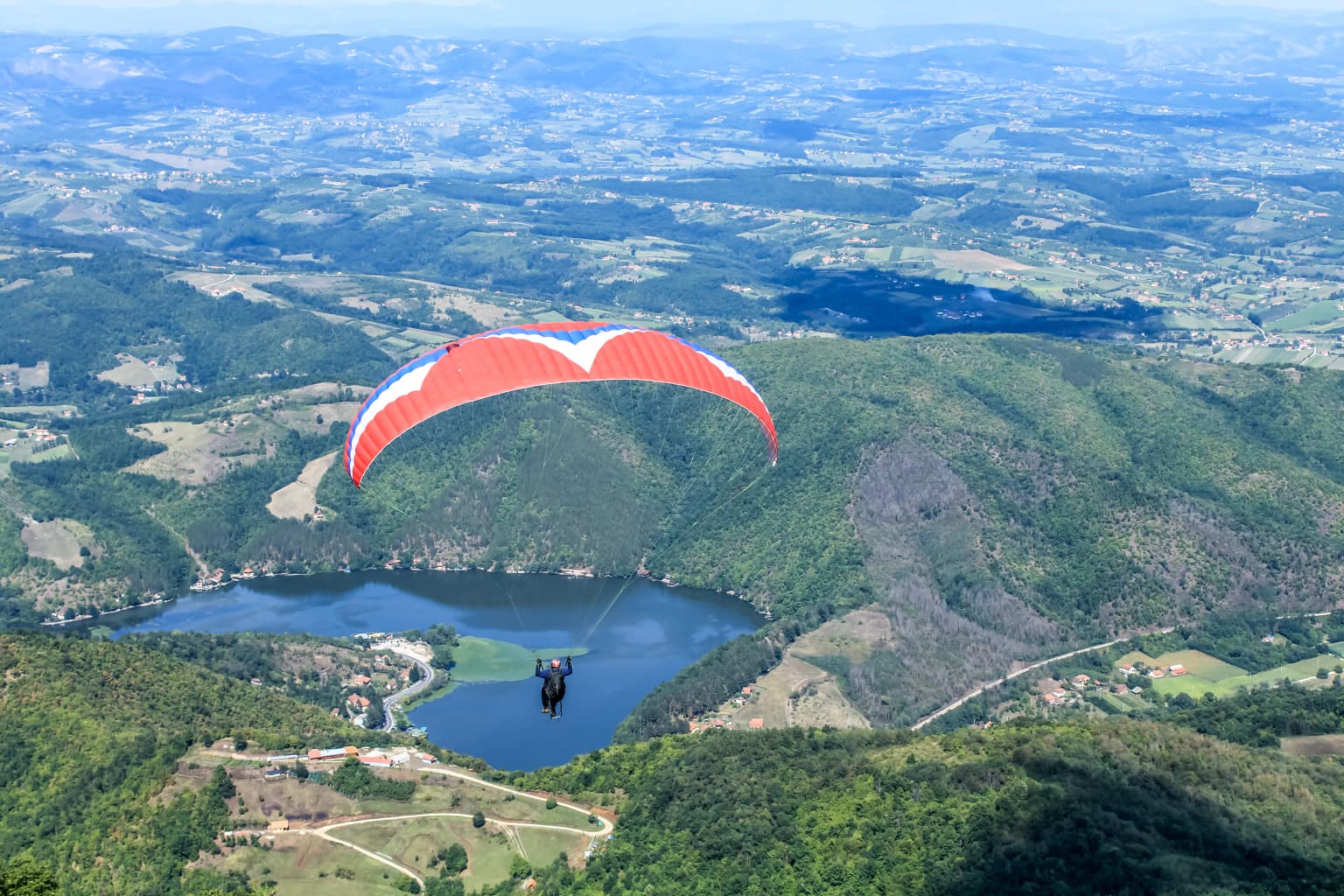 A red and white paraglider flying over a winding river valley surrounded by green hills and mountains