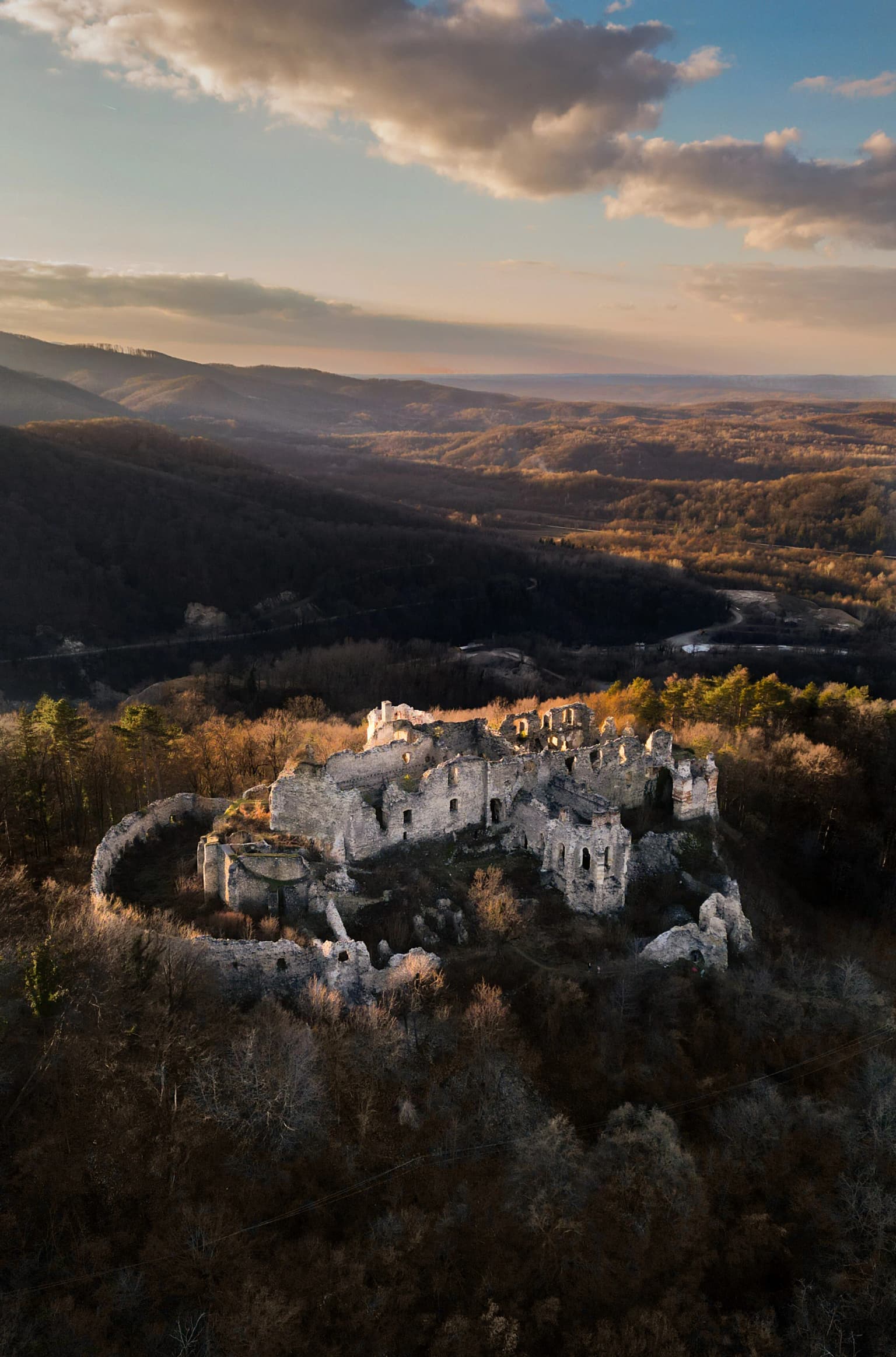 Aerial view of Ružica grad castle ruins atop a hill, surrounded by forested valleys and mountains under a partly cloudy sky