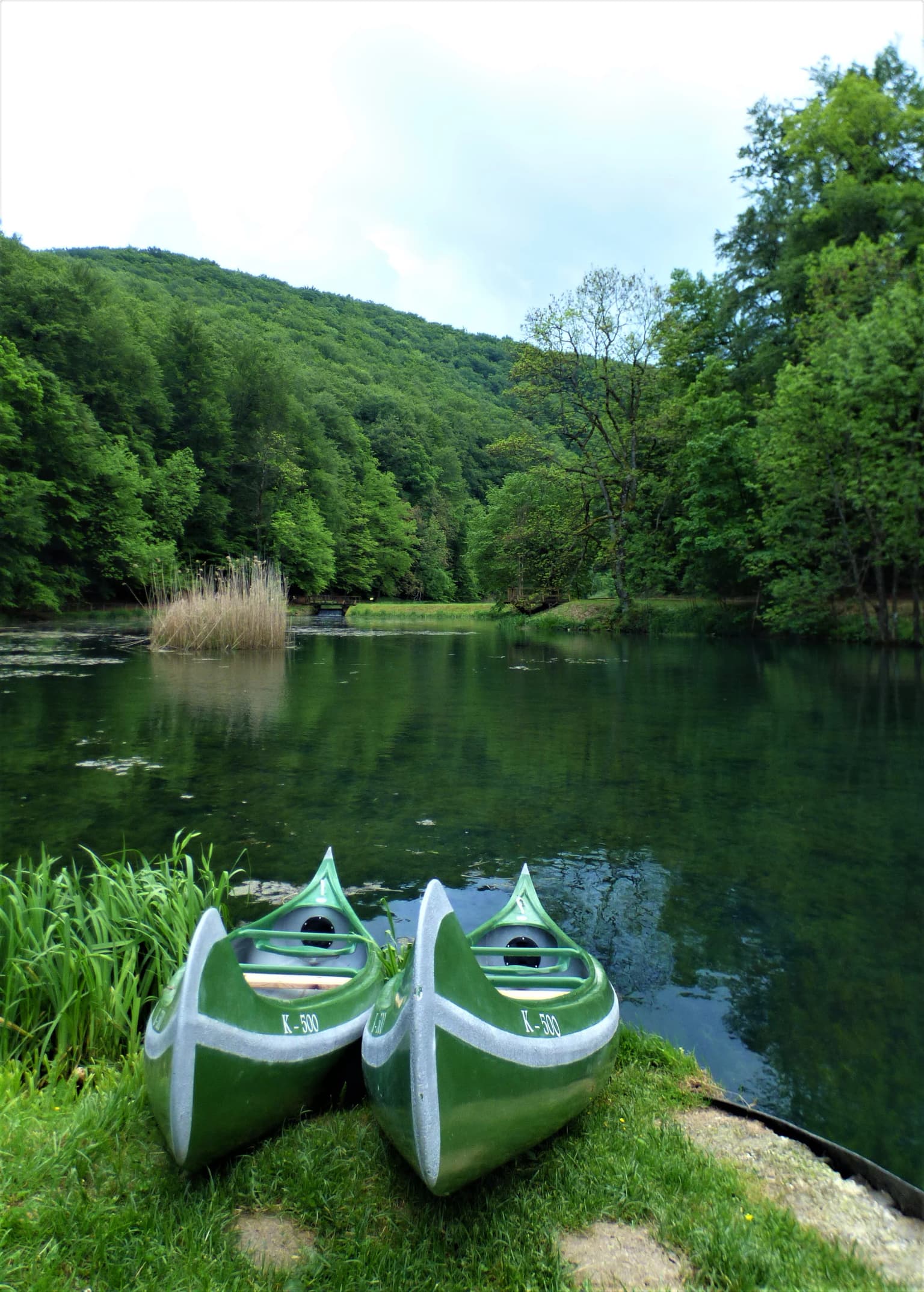 Two green kayaks with white trim resting on a grassy lakeshore, calm water reflecting surrounding trees and hills