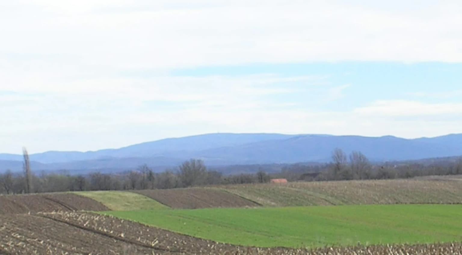 A landscape scene with plowed agricultural fields in the foreground, a green field, and the Papuk mountain range in the background under a partly cloudy sky