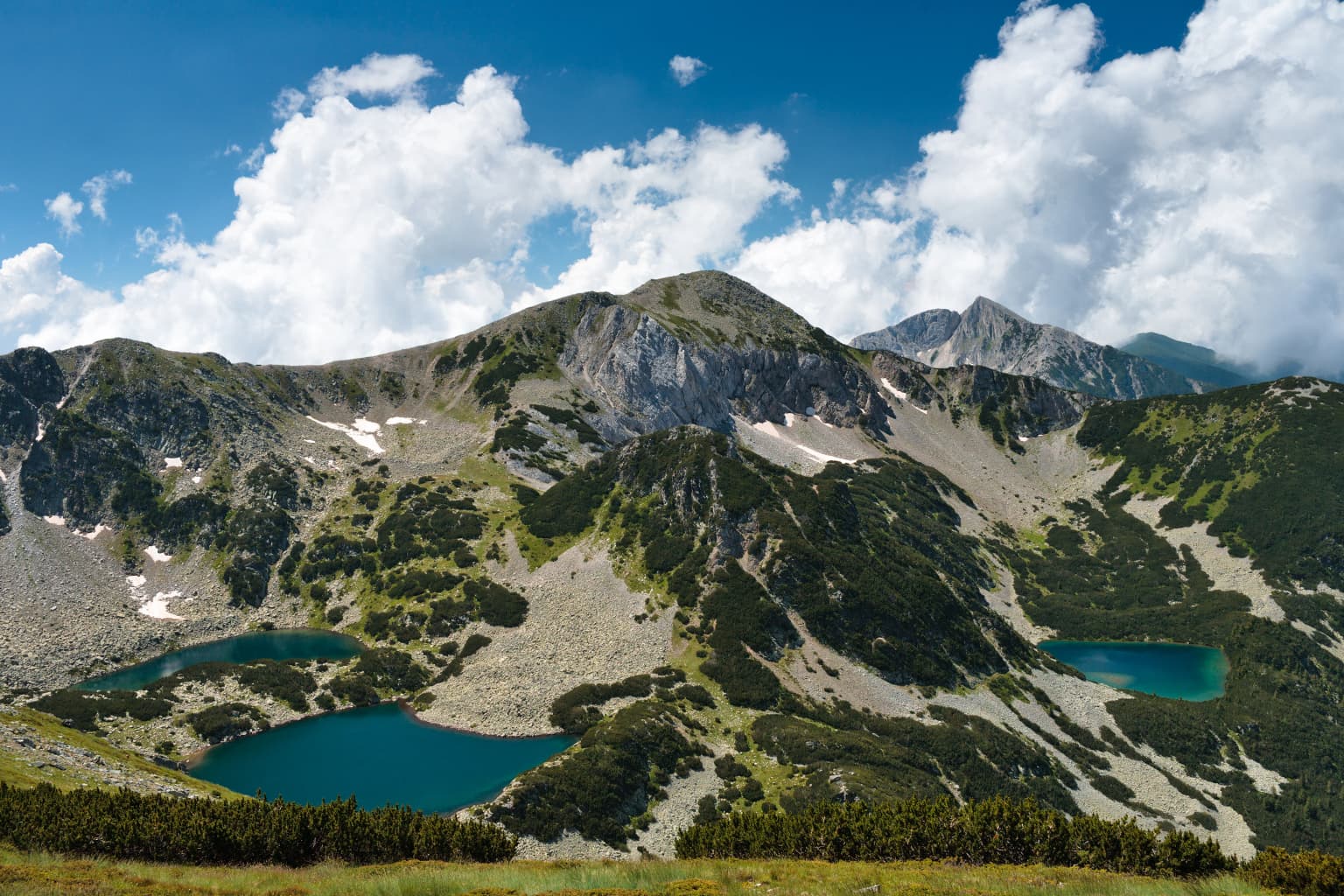 Mountain landscape with turquoise lakes nestled among rocky peaks and patches of snow under a blue sky with scattered clouds