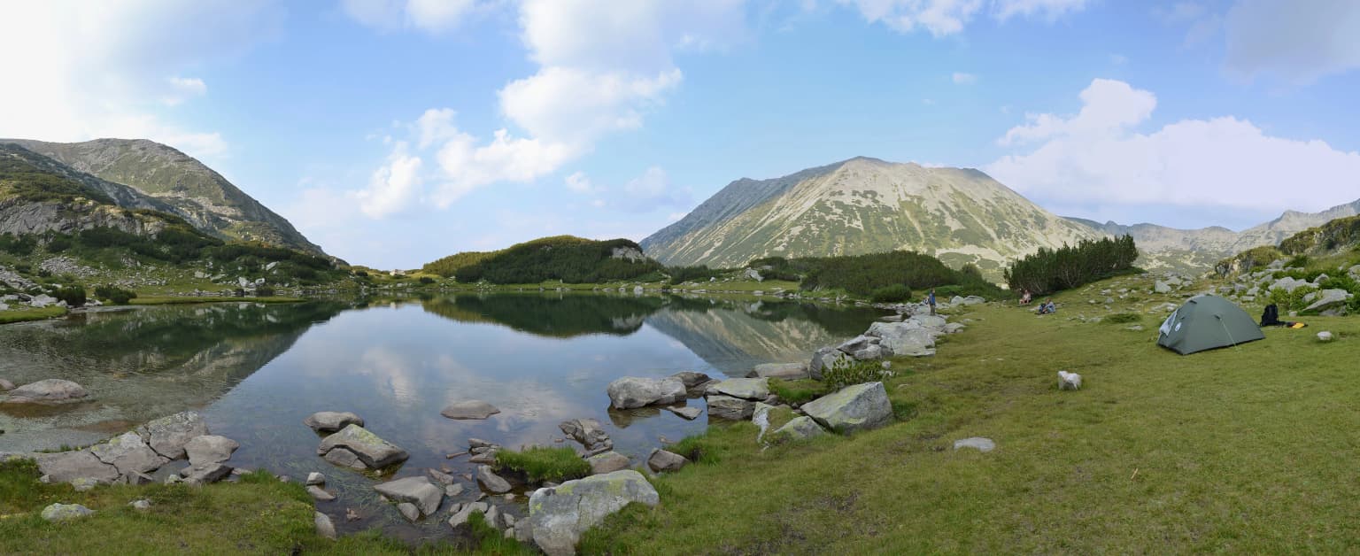 A calm lake reflecting mountain peaks under a partly cloudy sky, with rocky shoreline, green grassy terrain, and a tent visible on the right side