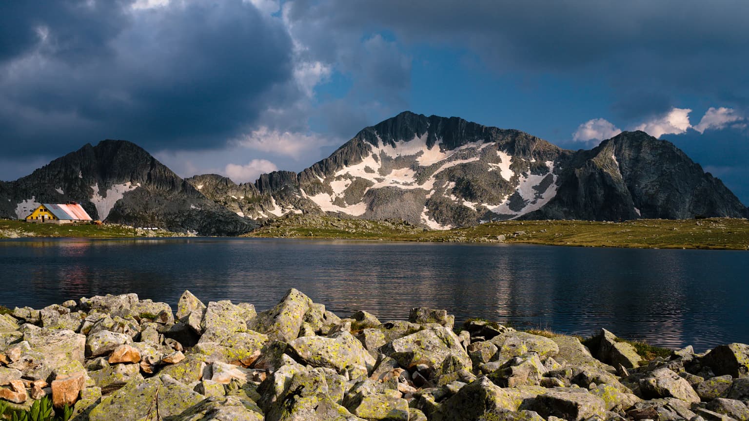 A lake with rocky shore in the foreground, mountains with snow patches in the background under a cloudy sky