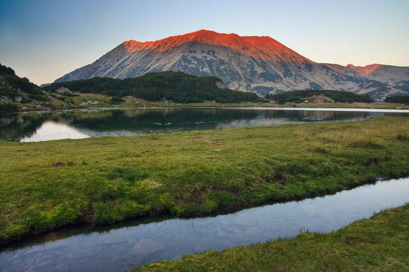 A mountain peak with a reddish hue reflecting in a calm lake, surrounded by grassy terrain