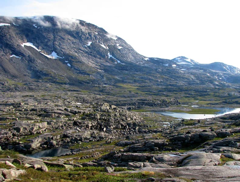 Rocky mountain landscape with patches of snow on peaks, a lake in the valley, and grassy areas