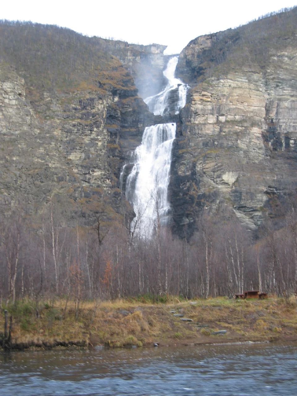 A tall waterfall flowing down rocky cliffs with a river in the foreground and sparse trees below.