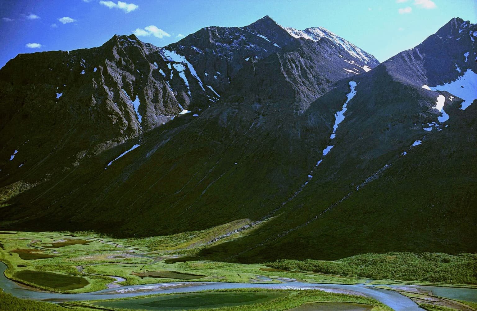 A panoramic view of Sarek National Park featuring rugged mountain peaks with patches of snow, a winding river through a green valley, and a clear blue sky