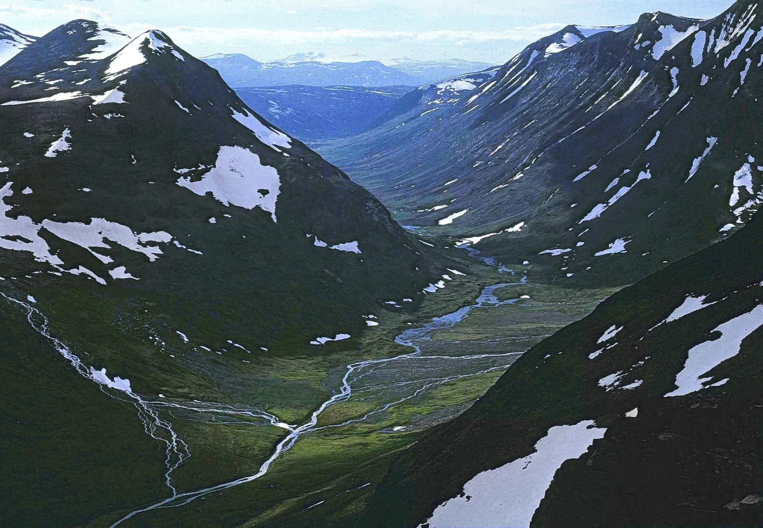 Aerial view of a mountainous valley with patches of snow, a winding river, and surrounding peaks