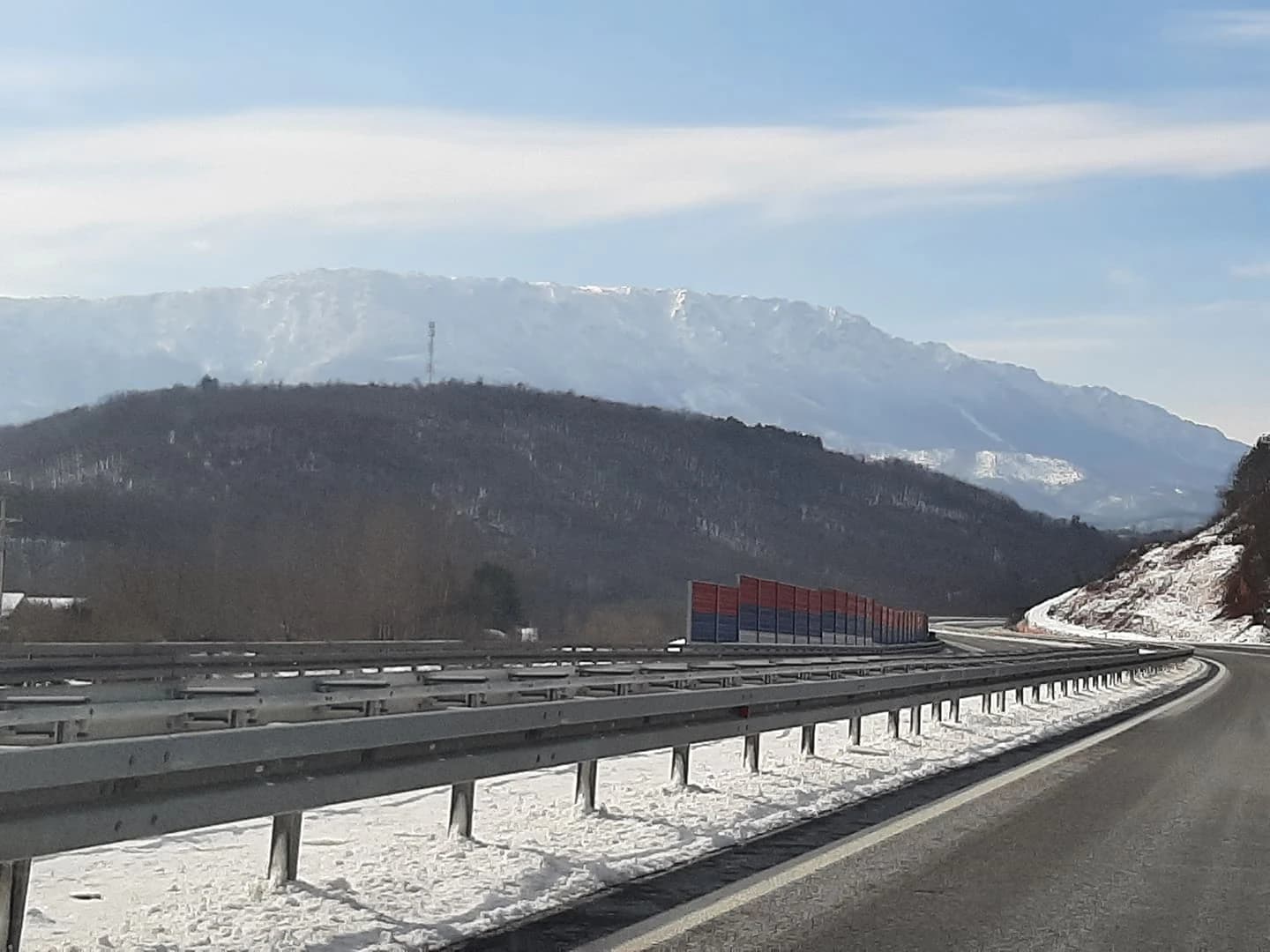 Highway with guardrails running through a mountainous landscape with snow-covered ground and distant mountain peaks under a clear sky