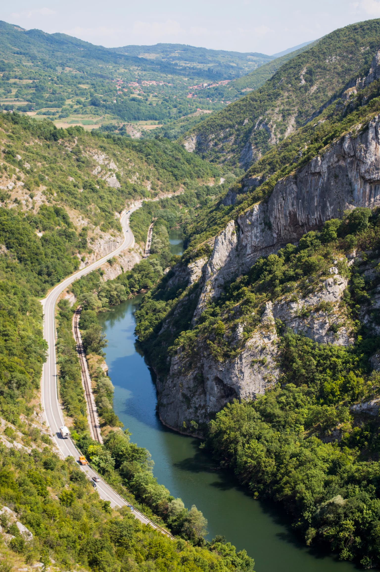 Aerial view of a river flowing through a gorge with steep limestone cliffs, a road with vehicles, and forested slopes