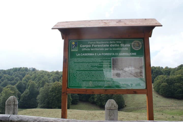 Green signboard with Italian text 'Parco Forestale dello Stato' and 'La Caserma e la Foresta di Gariglione' mounted on wooden posts, showing forested landscape in the background