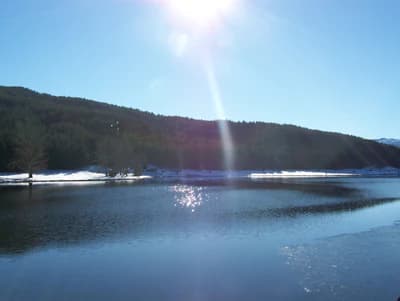 A calm lake reflecting sunlight with snow-covered mountains and forested hills under a clear blue sky