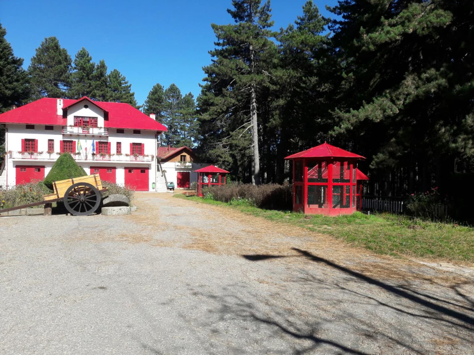 Red-roofed building with white walls and red shutters, wooden cart, and red gazebo on gravel path surrounded by pine trees