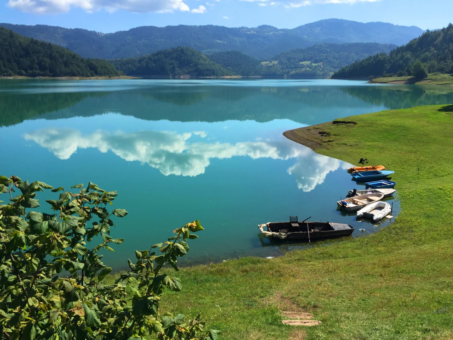 A scenic view of a turquoise lake reflecting clouds, with boats docked at a grassy shore and forested mountains in the background