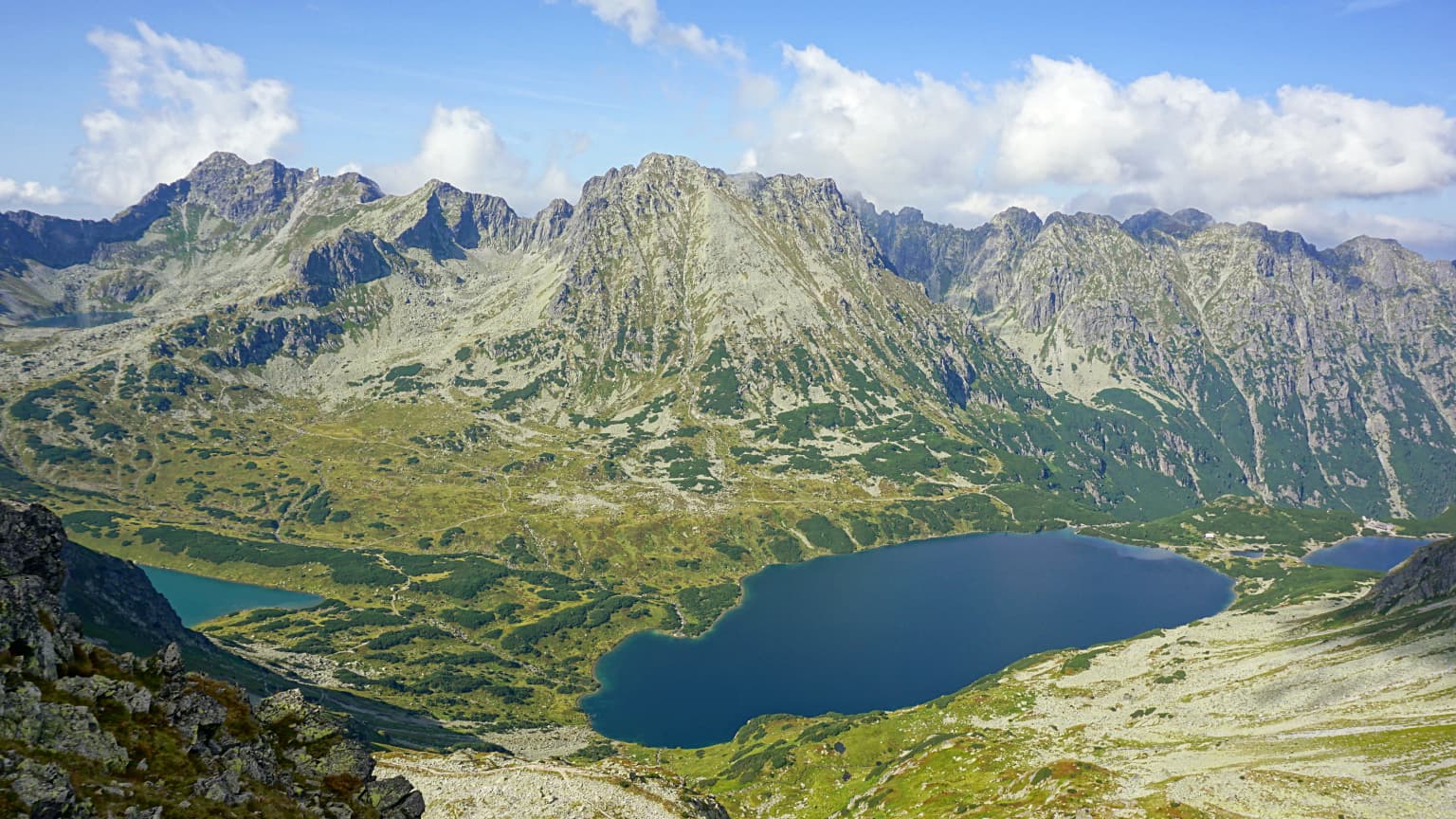 A panoramic alpine landscape featuring blue lakes nestled in mountain valleys with rocky peaks under a partly cloudy sky