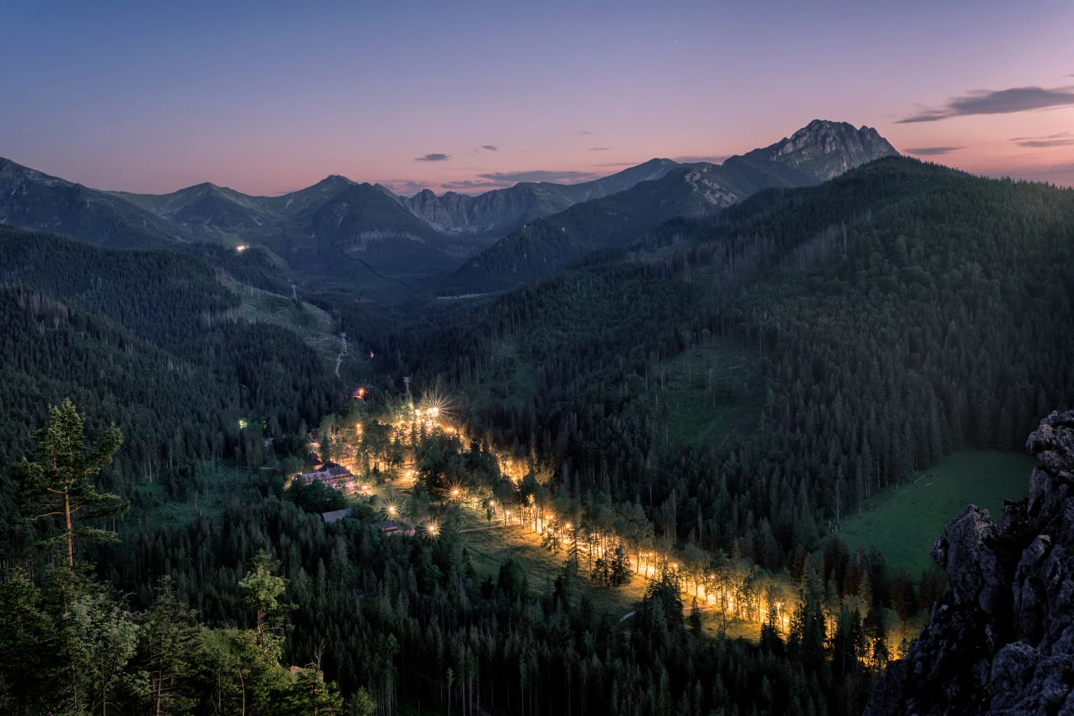 Mountain landscape at dusk with forested hills, illuminated valley path, and mountain peaks
