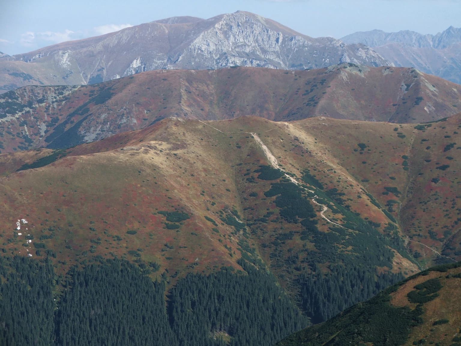 Wide mountain landscape showing multiple peaks with varied terrain, vegetation, and clear sky