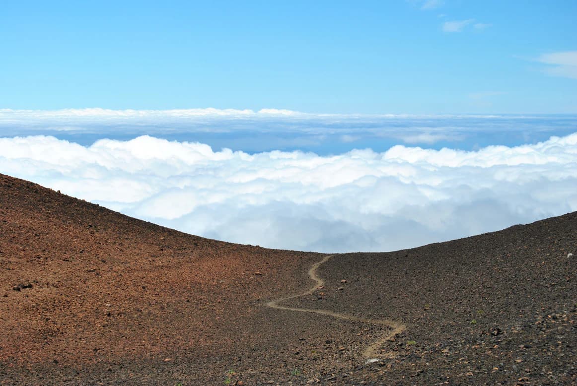 Winding dirt trail on rocky volcanic terrain with sea of clouds below under clear blue sky