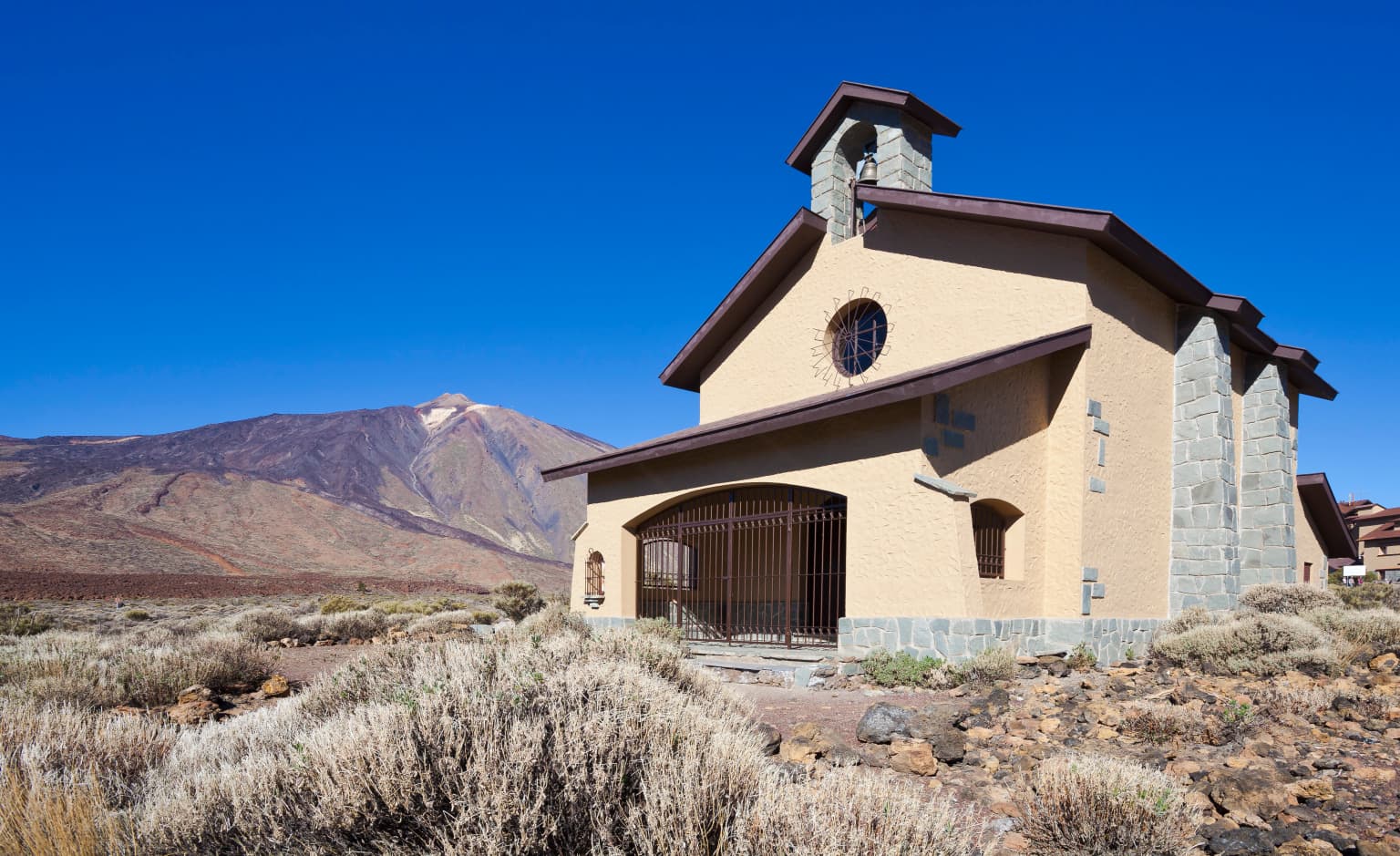 Small adobe-style building with stone accents and a bell tower in a volcanic landscape with Mount Teide visible on the horizon under a clear blue sky