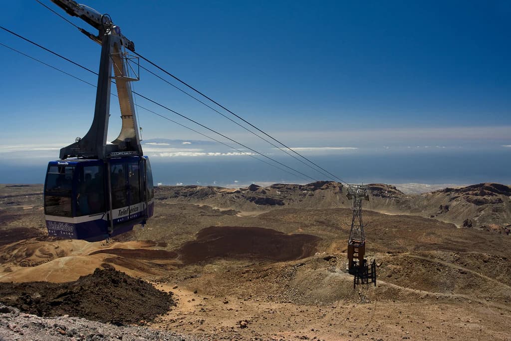 Cable car suspended on cables above barren volcanic landscape with mountain peaks under clear blue sky