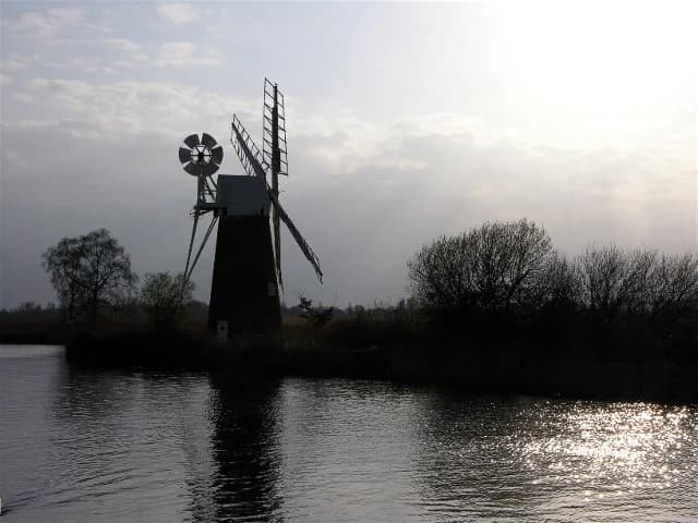 Windmill silhouette on a river with trees and a partly cloudy sky