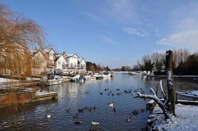 River with boats moored along a bank, snow-covered trees and houses in the background, ducks swimming in the water