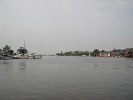 Calm water body with boats docked at shore and distant trees and buildings under overcast sky