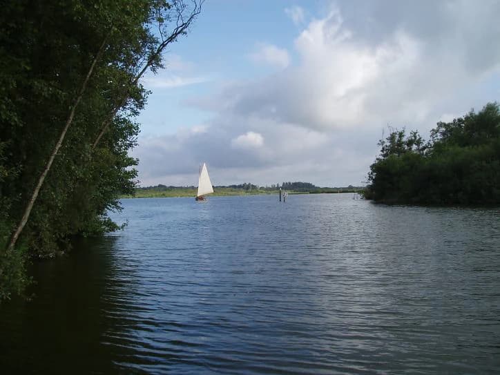 Calm water of Barton Broad with a white sailboat, trees on the left, and partly cloudy sky