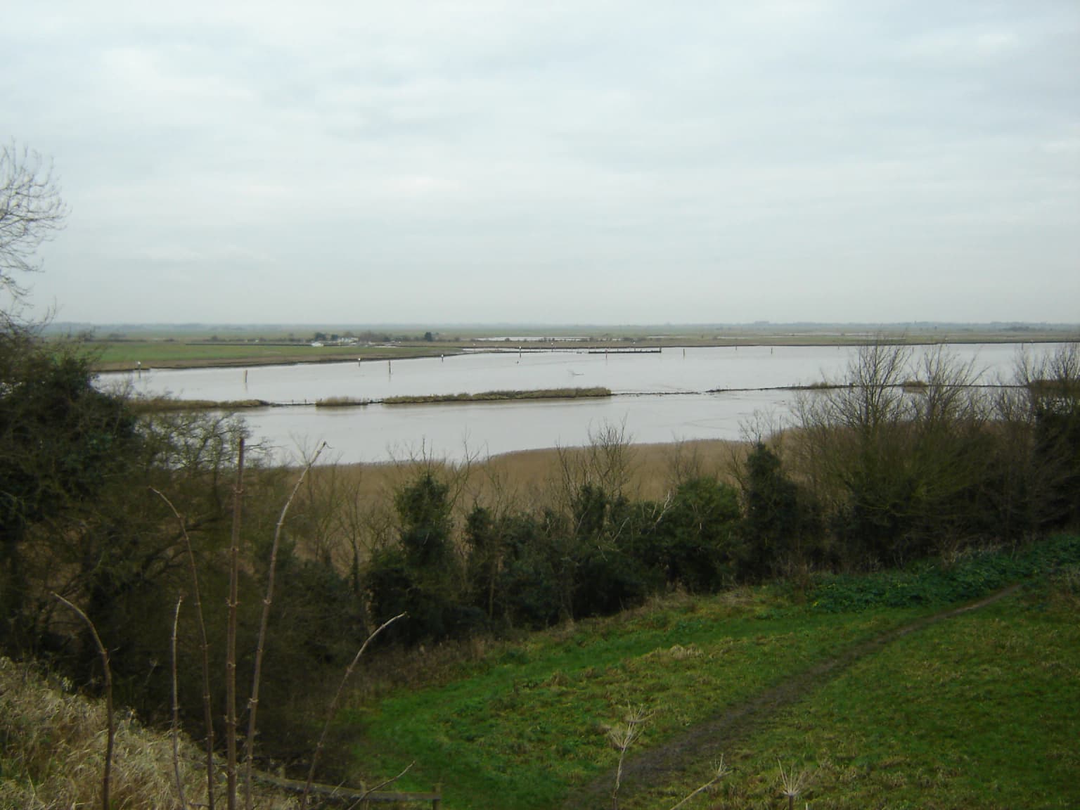 Wide landscape view of Breydon Water with a large body of water, grassy fields, and trees under an overcast sky.
