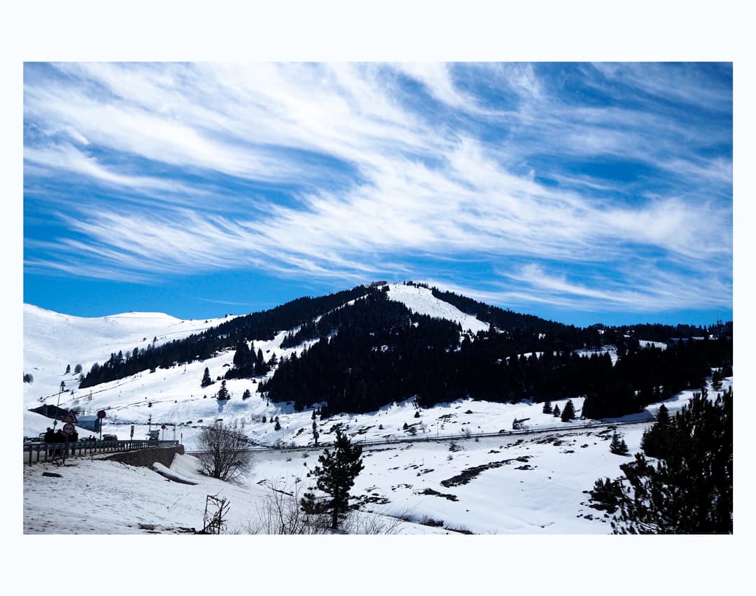 Snow-covered mountain slopes with a winding road, scattered trees, and a blue sky with wispy clouds