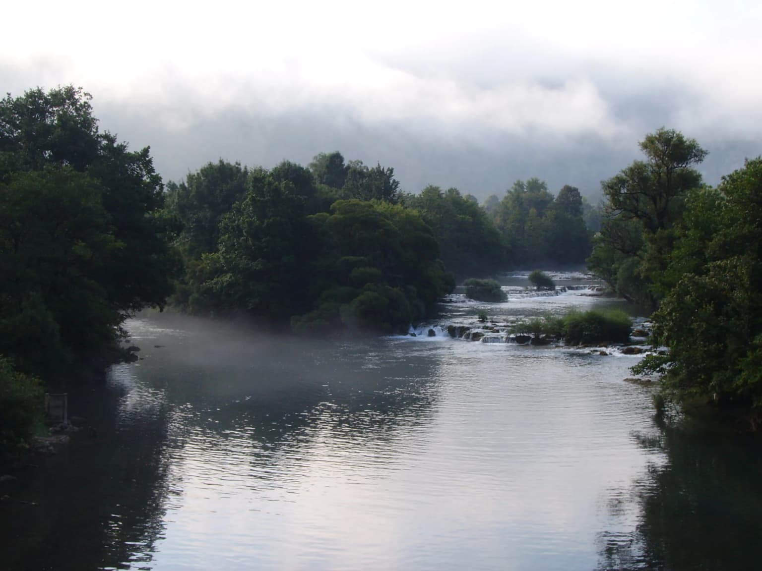 River confluence with mist rising over water, surrounded by dense green trees and a cloudy sky