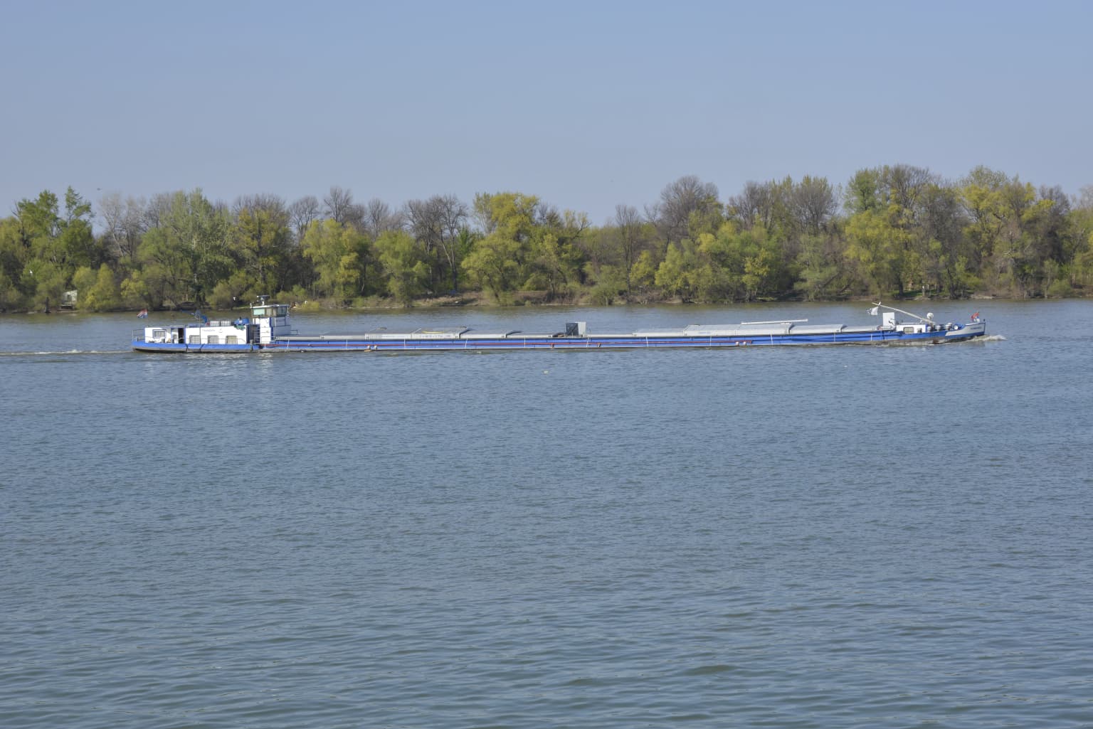 Cargo barge floating on a wide river with calm waters, forested shoreline and clear blue sky
