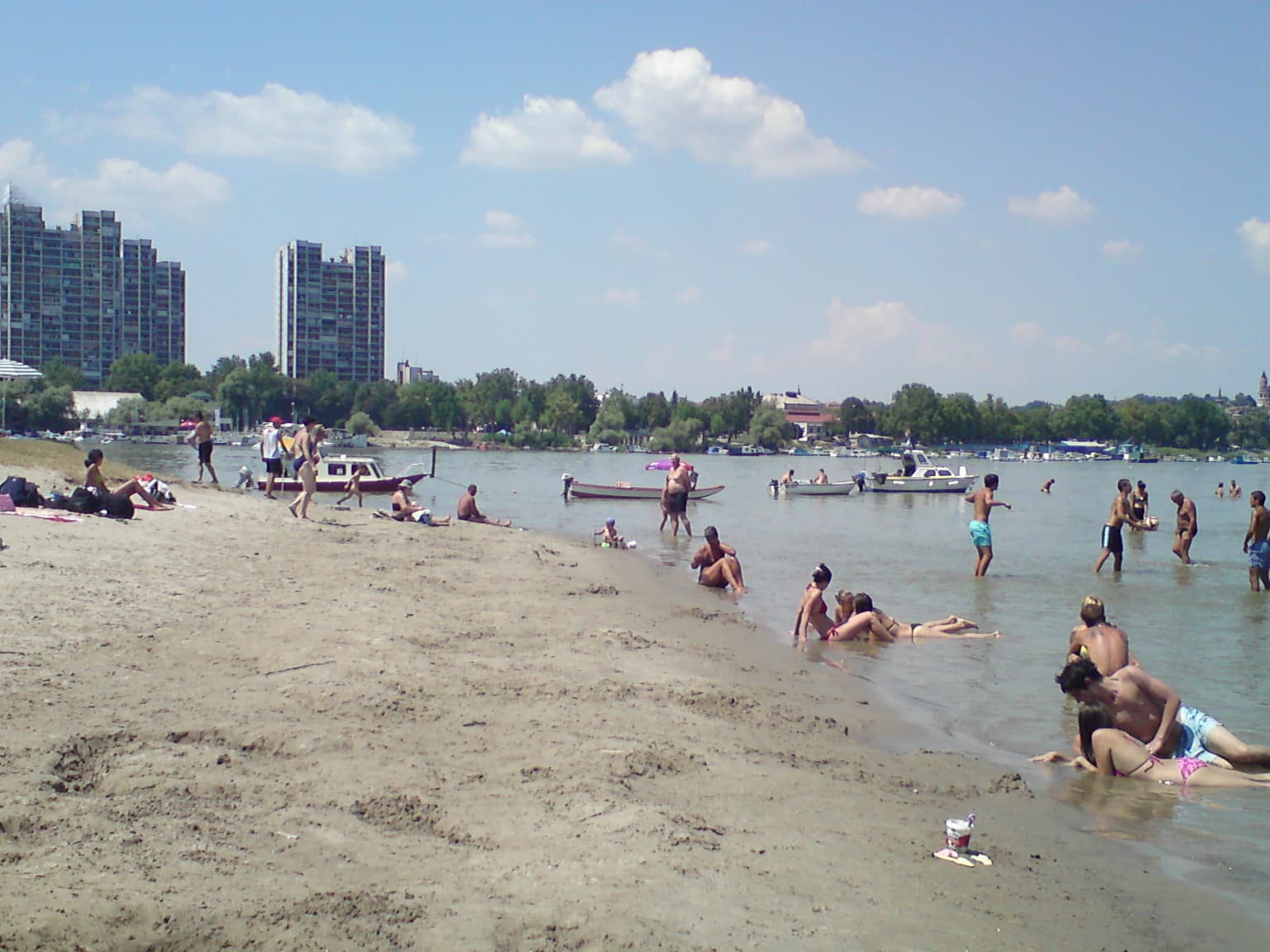 People swimming and relaxing on a sandy beach with boats on the Danube River, buildings visible in the background