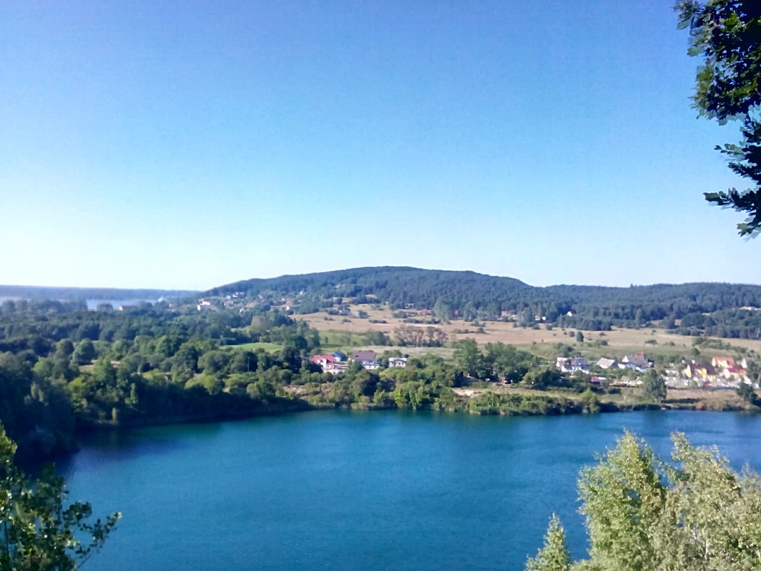 A panoramic view of a large blue lake surrounded by green forests and fields, with a hill in the background under a clear sky