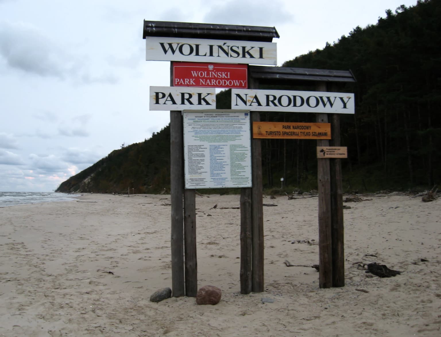 Wooden signpost with 'WOLIŃSKI PARK NARODOWY' text on sandy beach with forested hillside background