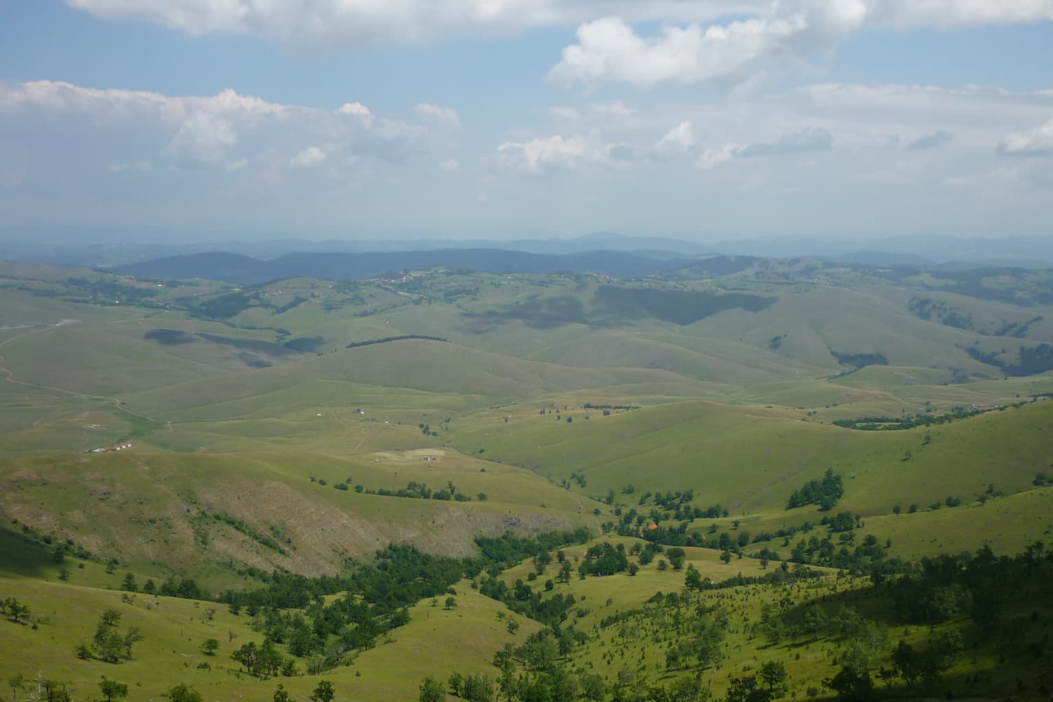 Wide panoramic view of rolling green hills and valleys with scattered trees under a partly cloudy sky