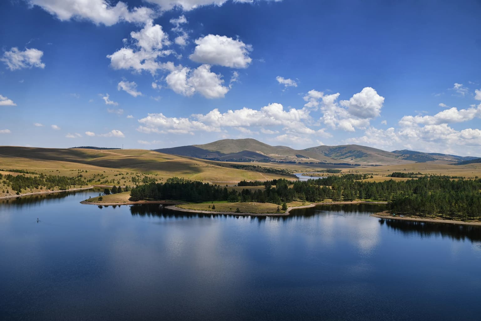 Landscape view of Ribnik Lake with clouds reflected in the water, surrounded by forested hills and mountains under a blue sky