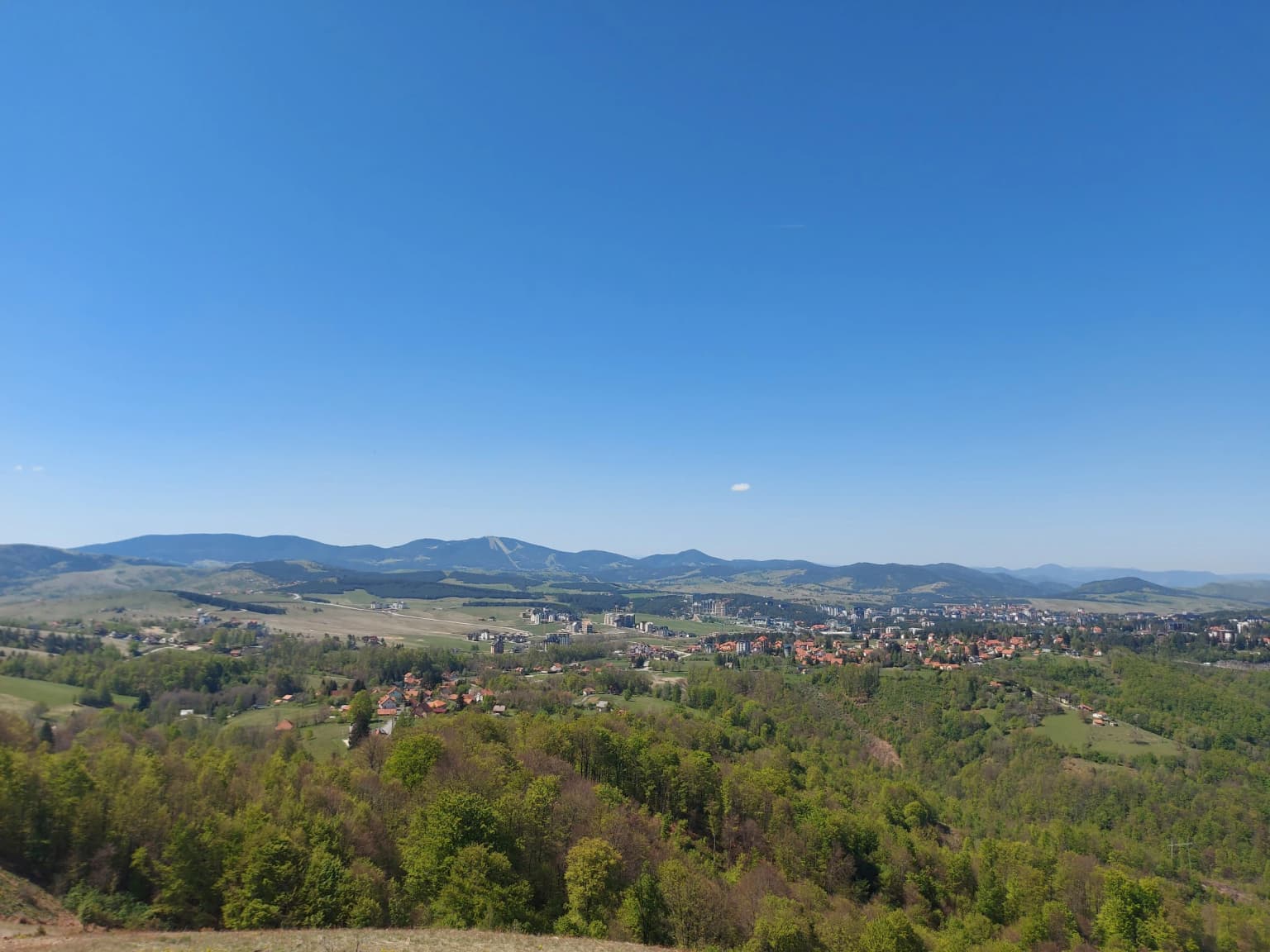 Wide landscape showing valley with town, green hills, and distant mountains under clear blue sky