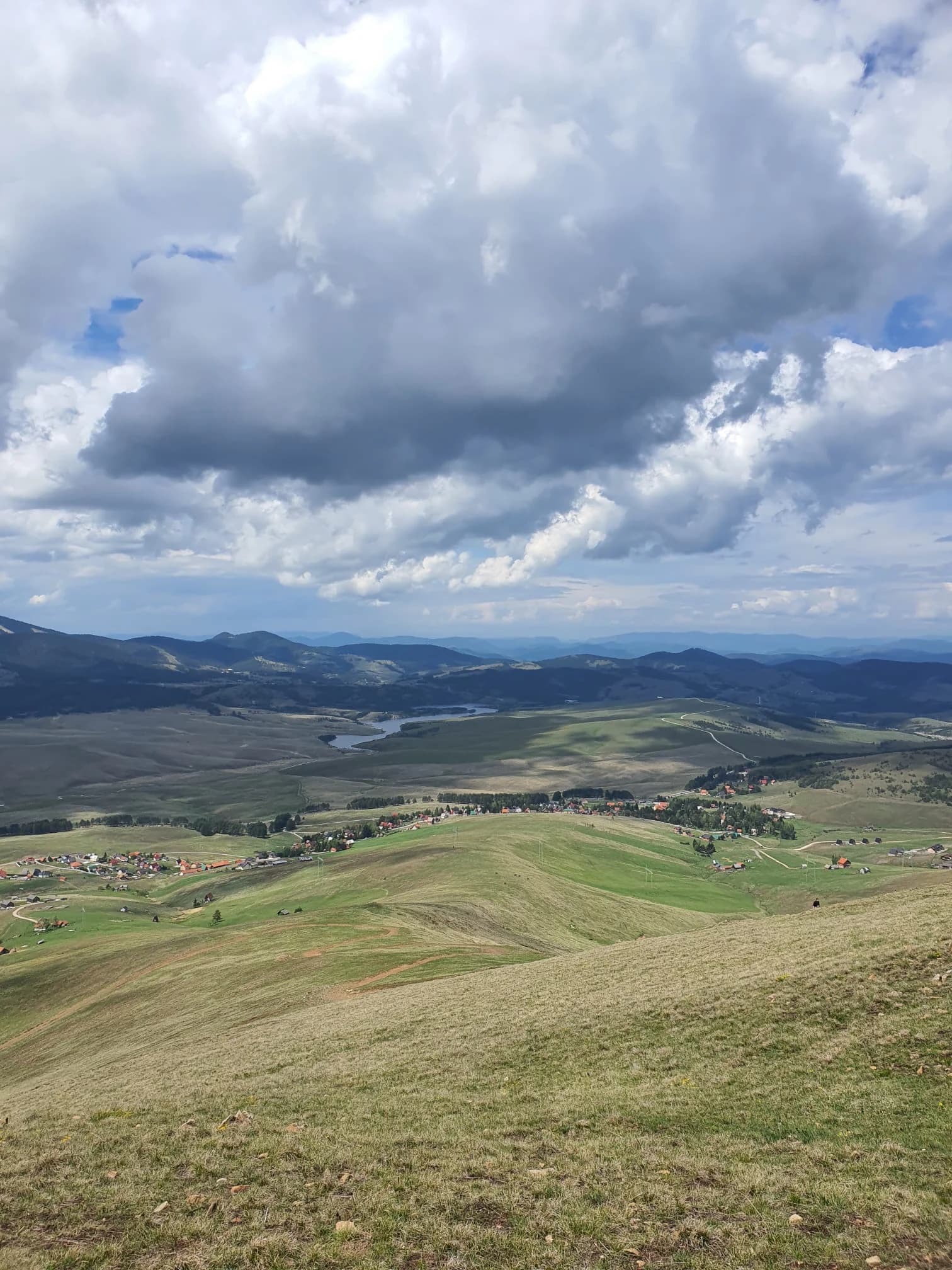 Wide landscape view from a mountain peak with rolling hills, a lake, distant mountains, scattered buildings, and partly cloudy sky
