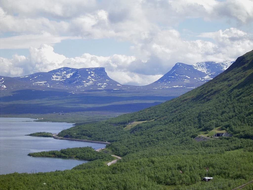 Landscape view of Lapporten valley with Torneträsk lake, snow-capped mountains, and dense green forest under a partly cloudy sky