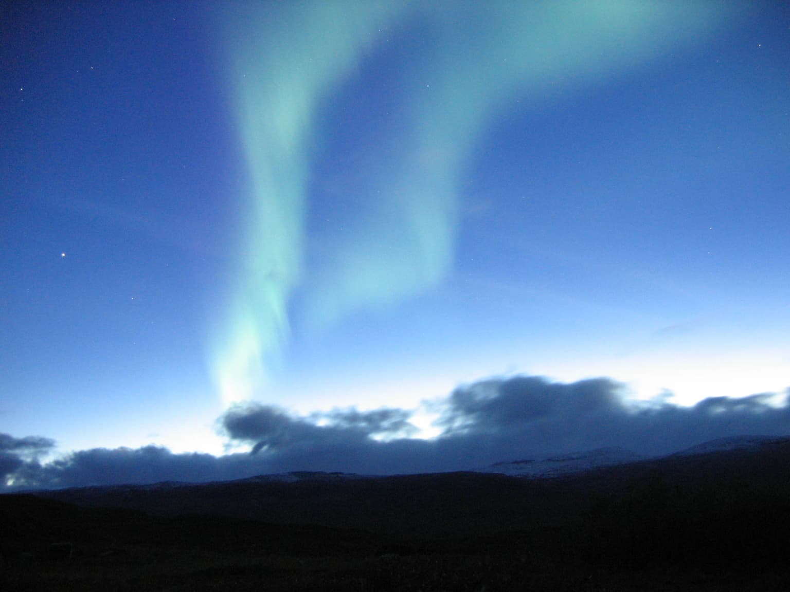 Northern lights displaying green and blue hues against a dark night sky with distant clouds and mountain silhouettes