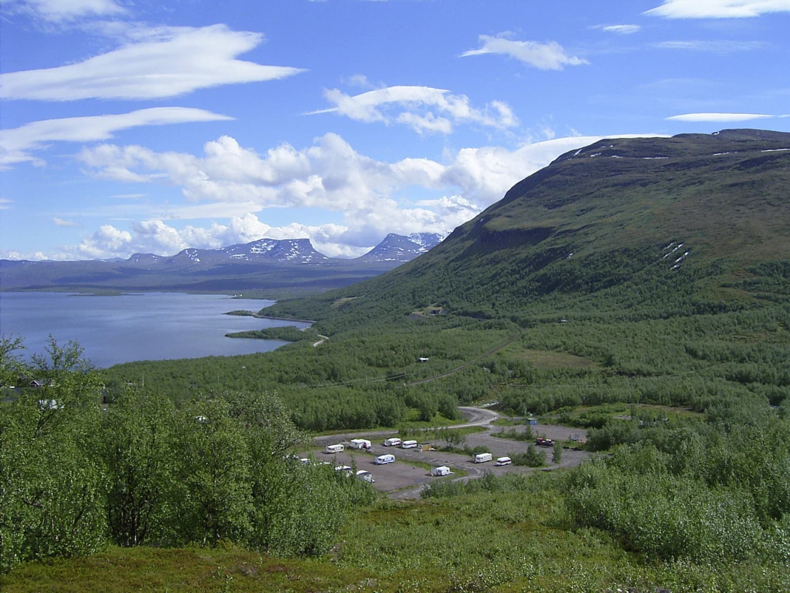 Panoramic landscape view showing a U-shaped valley, lake, mountainous terrain with green vegetation, and a parking area with vehicles under a partly cloudy sky