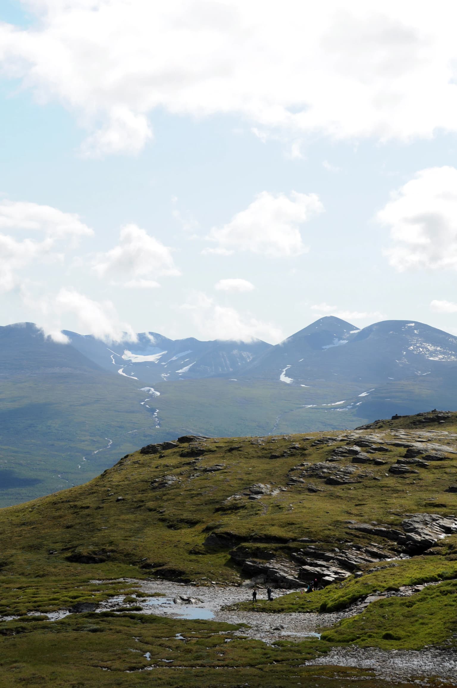 Grassy hillside with rocky outcrops, small stream, and distant snow-capped mountains under a partly cloudy sky