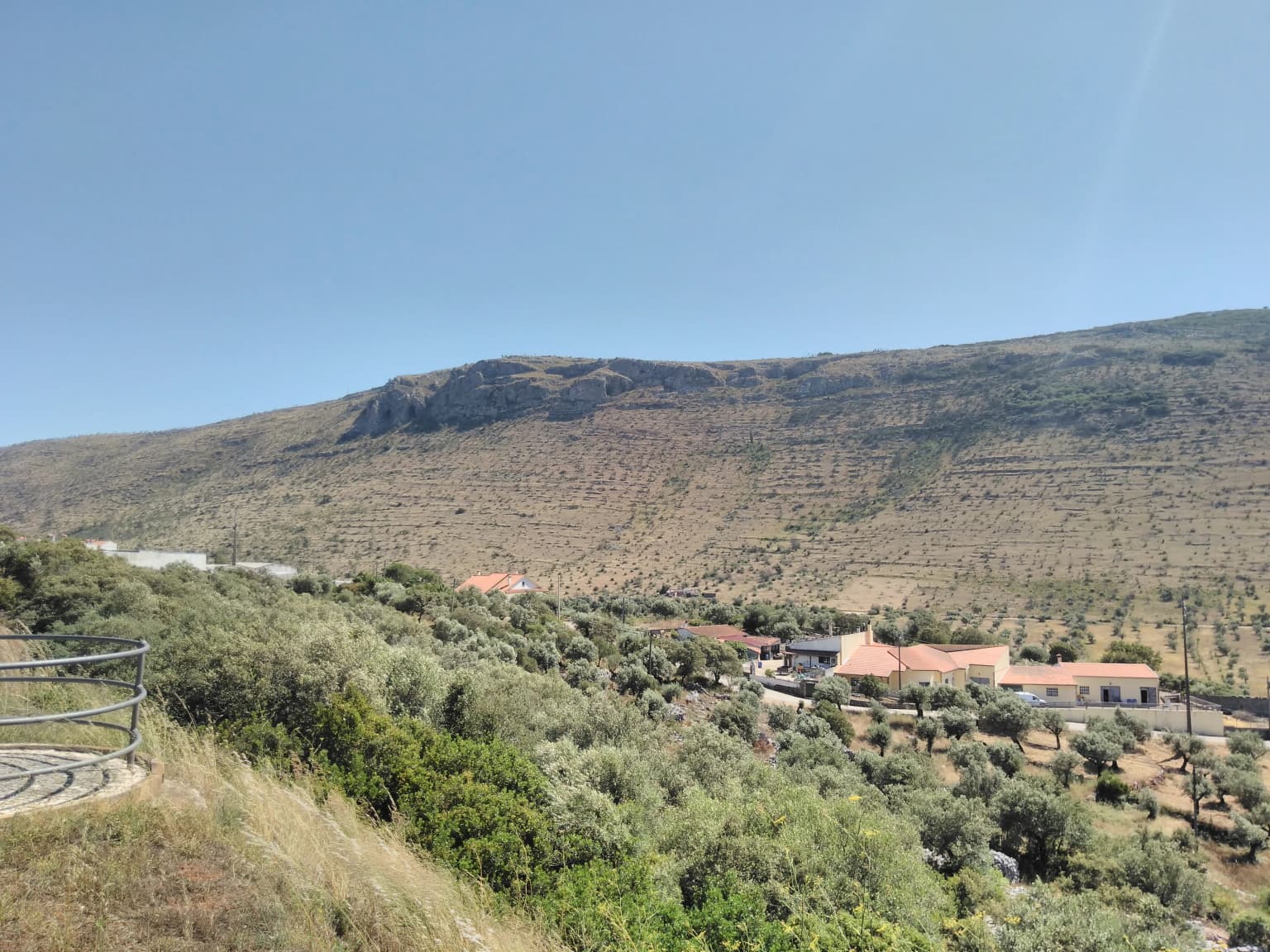 Wide landscape view of Serra de Santo António mountain range with a village, red-roofed houses, roads, and vegetation under clear blue sky