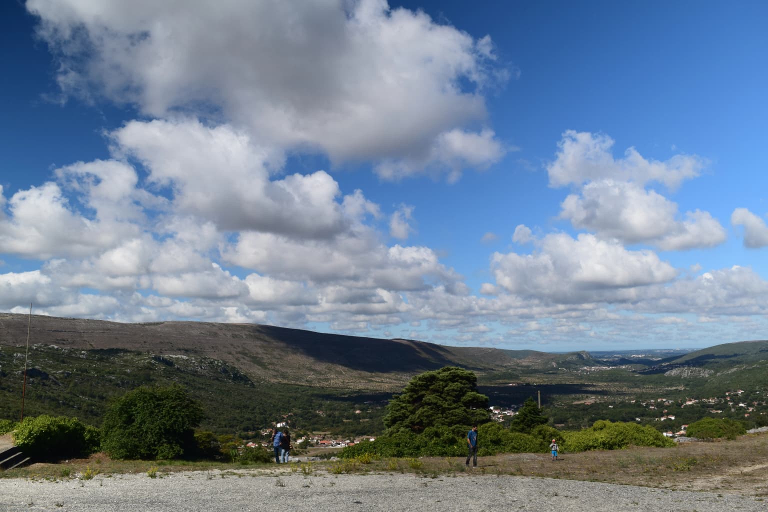 Wide landscape view of rolling hills and valleys under a blue sky with scattered clouds, featuring two people walking on a gravel path with trees and distant settlements