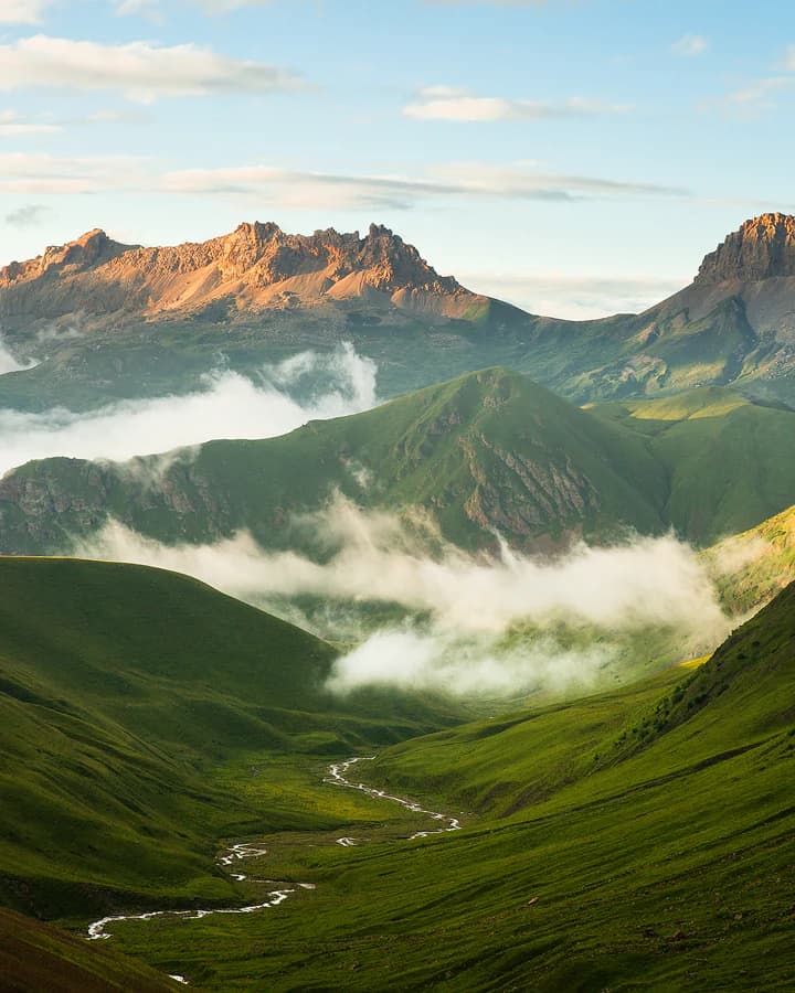 A wide view of a mountainous gorge with green slopes, a winding river, and low-lying clouds between rocky peaks